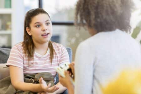 A teenage girl with brown hair in a ponytail, wearing a pink striped shirt, sits on a sofa and talks to a woman holding a notepad, seen from behind. The setting appears to be a casual, well-lit counselling or therapy session.