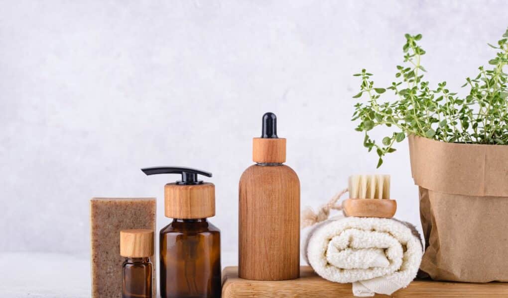 A set of natural skincare items on a wooden tray: amber glass bottles with wooden lids, a bar of soap, a dropper bottle, a pump bottle, a rolled white towel, a brush, and a paper-wrapped potted plant, all against a light background.