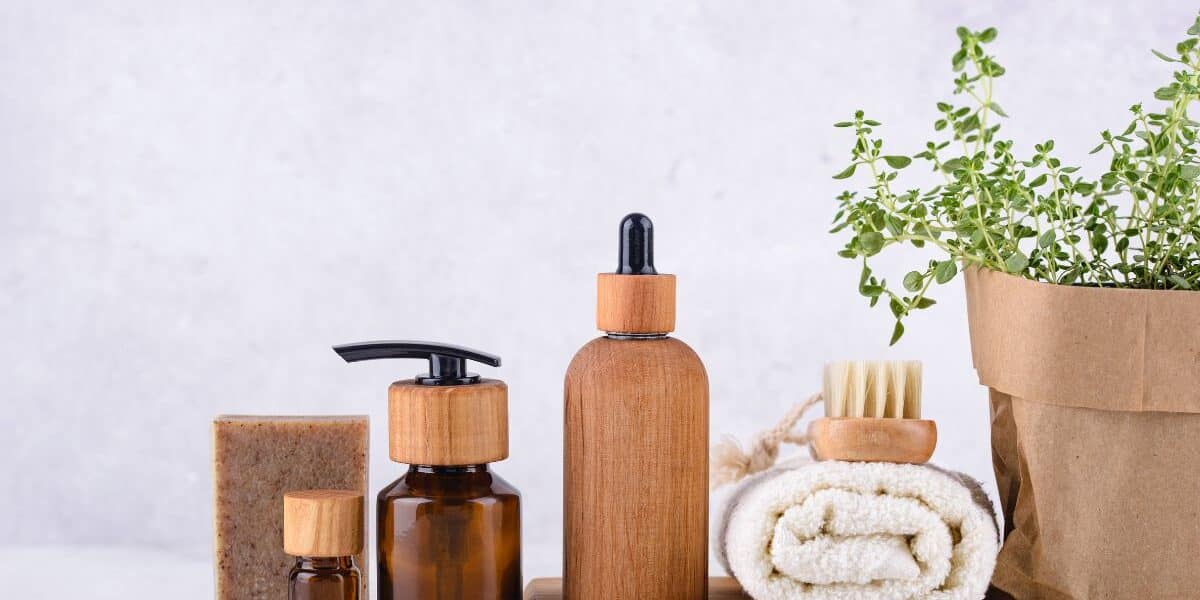 A set of natural skincare items on a wooden tray: amber glass bottles with wooden lids, a bar of soap, a dropper bottle, a pump bottle, a rolled white towel, a brush, and a paper-wrapped potted plant, all against a light background.