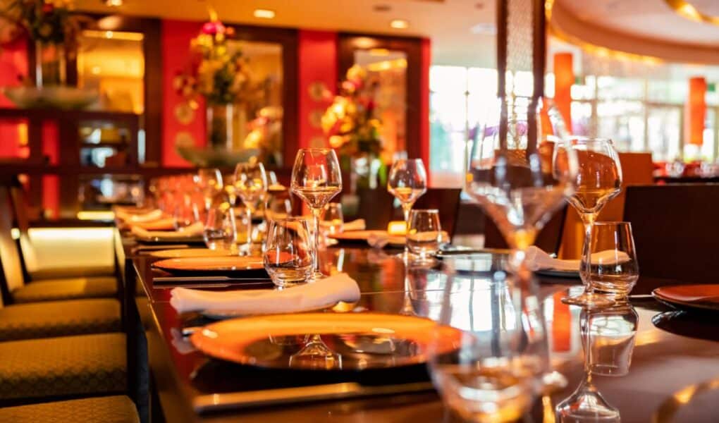 A close-up view of a stylish restaurant table set for dining, featuring orange plates, neatly rolled white serviettes, clear wine glasses, and water glasses. The background shows vibrant red walls, warm lighting, and decorative flower arrangements.