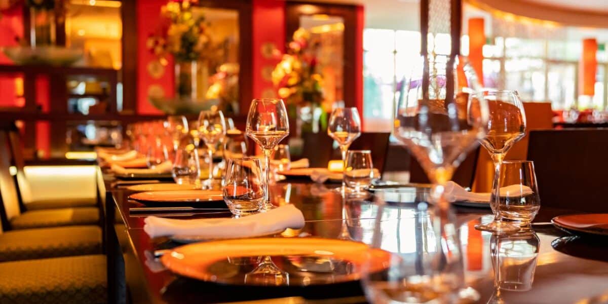 A close-up view of a stylish restaurant table set for dining, featuring orange plates, neatly rolled white serviettes, clear wine glasses, and water glasses. The background shows vibrant red walls, warm lighting, and decorative flower arrangements.
