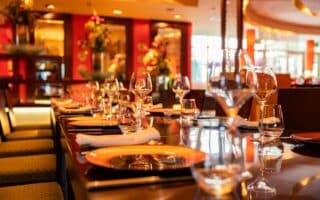 A close-up view of a stylish restaurant table set for dining, featuring orange plates, neatly rolled white serviettes, clear wine glasses, and water glasses. The background shows vibrant red walls, warm lighting, and decorative flower arrangements.