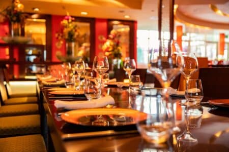 A close-up view of a stylish restaurant table set for dining, featuring orange plates, neatly rolled white serviettes, clear wine glasses, and water glasses. The background shows vibrant red walls, warm lighting, and decorative flower arrangements.