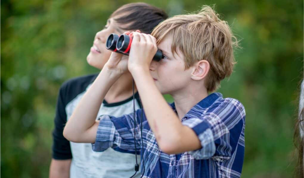 Two boys stand outdoors surrounded by greenery. The boy in front, wearing a checked shirt, is looking through black and red binoculars. The boy behind him, in a black and white shirt, looks up and smiles. Both appear curious and engaged with their surroundings.