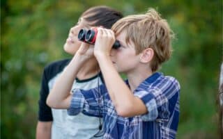 Two boys stand outdoors surrounded by greenery. The boy in front, wearing a checked shirt, is looking through black and red binoculars. The boy behind him, in a black and white shirt, looks up and smiles. Both appear curious and engaged with their surroundings.