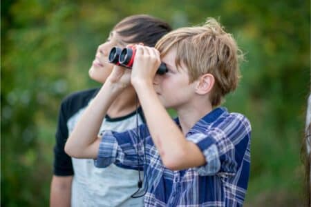 Two boys stand outdoors surrounded by greenery. The boy in front, wearing a checked shirt, is looking through black and red binoculars. The boy behind him, in a black and white shirt, looks up and smiles. Both appear curious and engaged with their surroundings.