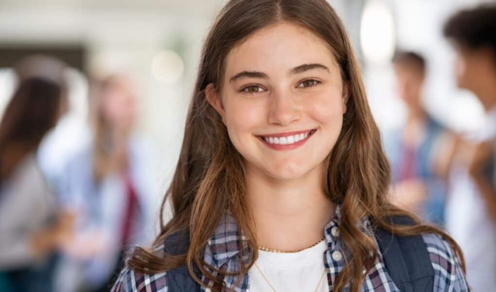 A teenage girl with long brown hair, wearing a checked shirt over a white top and a rucksack, smiles at the camera. The background shows a blurred group of students in a bright indoor setting, suggesting a school or college environment.