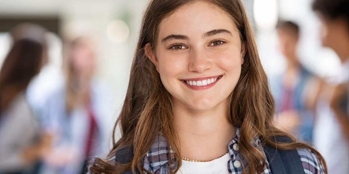 A teenage girl with long brown hair, wearing a checked shirt over a white top and a rucksack, smiles at the camera. The background shows a blurred group of students in a bright indoor setting, suggesting a school or college environment.