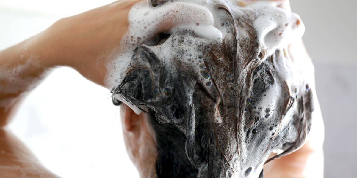 A person is washing their hair, seen from behind. Their hands are massaging shampoo into wet, dark hair, creating thick, white lather and bubbles. The background is softly blurred, focussing attention on the hair and foam.
