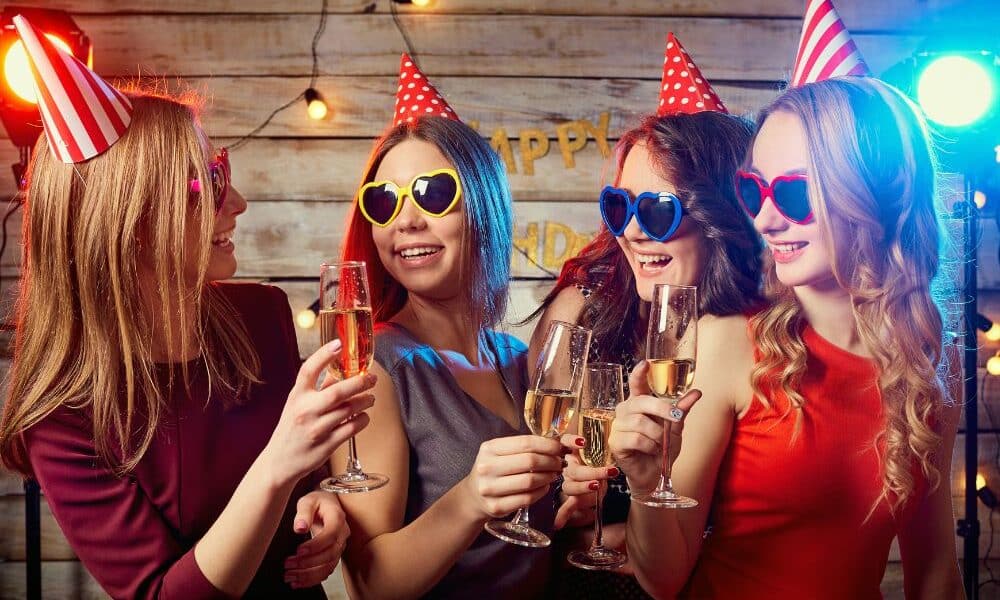 Four women wearing party hats and heart-shaped sunglasses smile and toast with champagne glasses. They stand closely together in front of a wooden wall decorated with fairy lights and a “Happy Birthday” banner, with colourful party lights in the background.