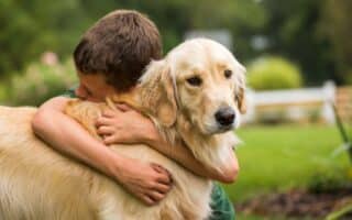 A young boy with short brown hair hugs a golden retriever tightly outdoors. The dog looks ahead with a calm expression whilst the boy's face is pressed against its neck. Green grass and blurred plants fill the background.