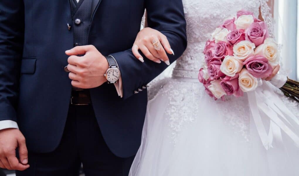 A bride in a white lace gown holds a bouquet of pink and cream roses, linking arms with a groom in a dark suit and watch. Their faces are not visible; the focus is on their torsos, clasped arms, and the bouquet.