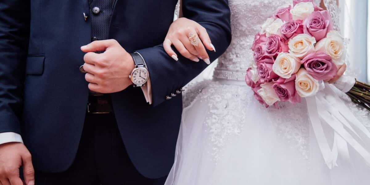 A bride in a white lace gown holds a bouquet of pink and cream roses, linking arms with a groom in a dark suit and watch. Their faces are not visible; the focus is on their torsos, clasped arms, and the bouquet.