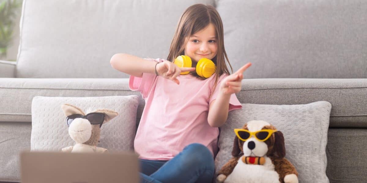 A young girl sits cross-legged on the floor in front of a grey sofa, wearing headphones around her neck and making peace signs with both hands. Two soft toys with sunglasses are beside her, and a laptop sits open in the foreground.