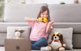 A young girl sits cross-legged on the floor in front of a grey sofa, wearing headphones around her neck and making peace signs with both hands. Two soft toys with sunglasses are beside her, and a laptop sits open in the foreground.