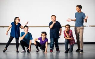 Six children stand and crouch in a row on a stage, striking expressive poses as if acting or performing. They wear casual clothing in various colours, and the background is a plain, light-coloured wall. The mood is energetic and playful.