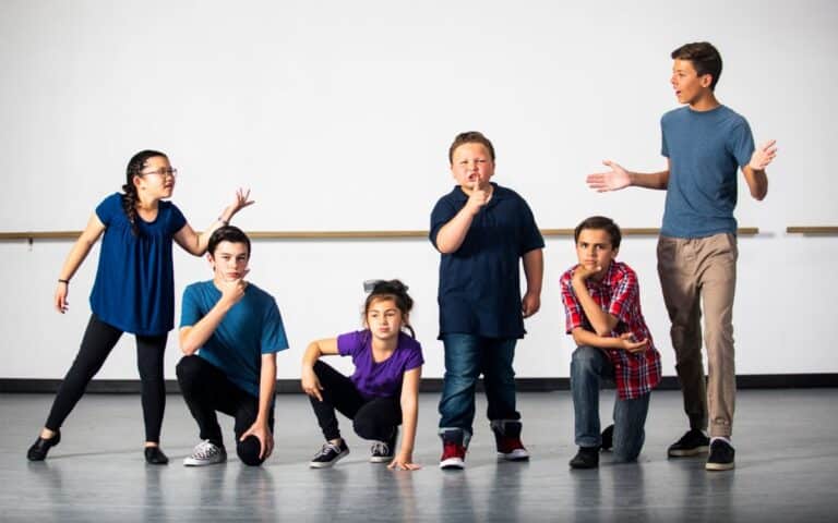 Six children stand and crouch in a row on a stage, striking expressive poses as if acting or performing. They wear casual clothing in various colours, and the background is a plain, light-coloured wall. The mood is energetic and playful.