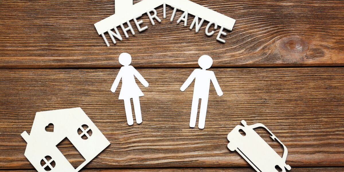 Cut-out figures of a man and woman stand under a roof labelled "INHERITANCE." Below them are paper shapes of a house and a car, all arranged on a wooden surface, symbolising dividing inherited assets like property and vehicles.