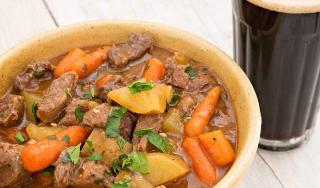 A bowl of hearty beef stew with chunks of beef, potatoes, and whole baby carrots in a rich brown broth, garnished with chopped parsley. Next to the bowl is a glass of dark beer with a creamy head, set on a light wooden surface.