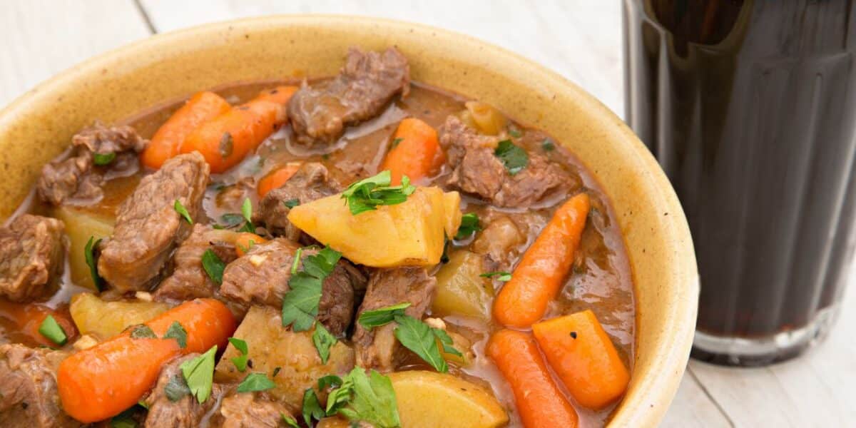 A bowl of hearty beef stew with chunks of beef, potatoes, and whole baby carrots in a rich brown broth, garnished with chopped parsley. Next to the bowl is a glass of dark beer with a creamy head, set on a light wooden surface.