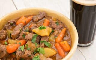 A bowl of hearty beef stew with chunks of beef, potatoes, and whole baby carrots in a rich brown broth, garnished with chopped parsley. Next to the bowl is a glass of dark beer with a creamy head, set on a light wooden surface.