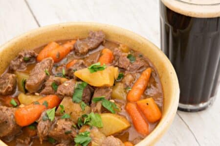 A bowl of hearty beef stew with chunks of beef, potatoes, and whole baby carrots in a rich brown broth, garnished with chopped parsley. Next to the bowl is a glass of dark beer with a creamy head, set on a light wooden surface.