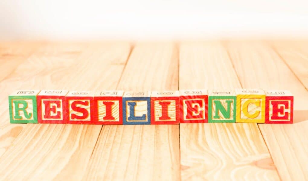 Colourful wooden alphabet blocks spell "RESILIENCE" on a light wooden surface. The blocks are arranged in a neat row and each letter is on a different coloured background, with sunlight casting soft shadows across the scene.