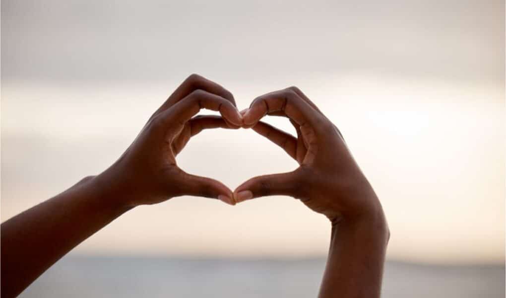 Two hands are held up against a blurred, bright sky, forming the shape of a heart with the fingers. The background is soft and out of focus, emphasising the hands and the heart gesture in the foreground.