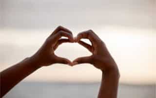 Two hands are held up against a blurred, bright sky, forming the shape of a heart with the fingers. The background is soft and out of focus, emphasising the hands and the heart gesture in the foreground.
