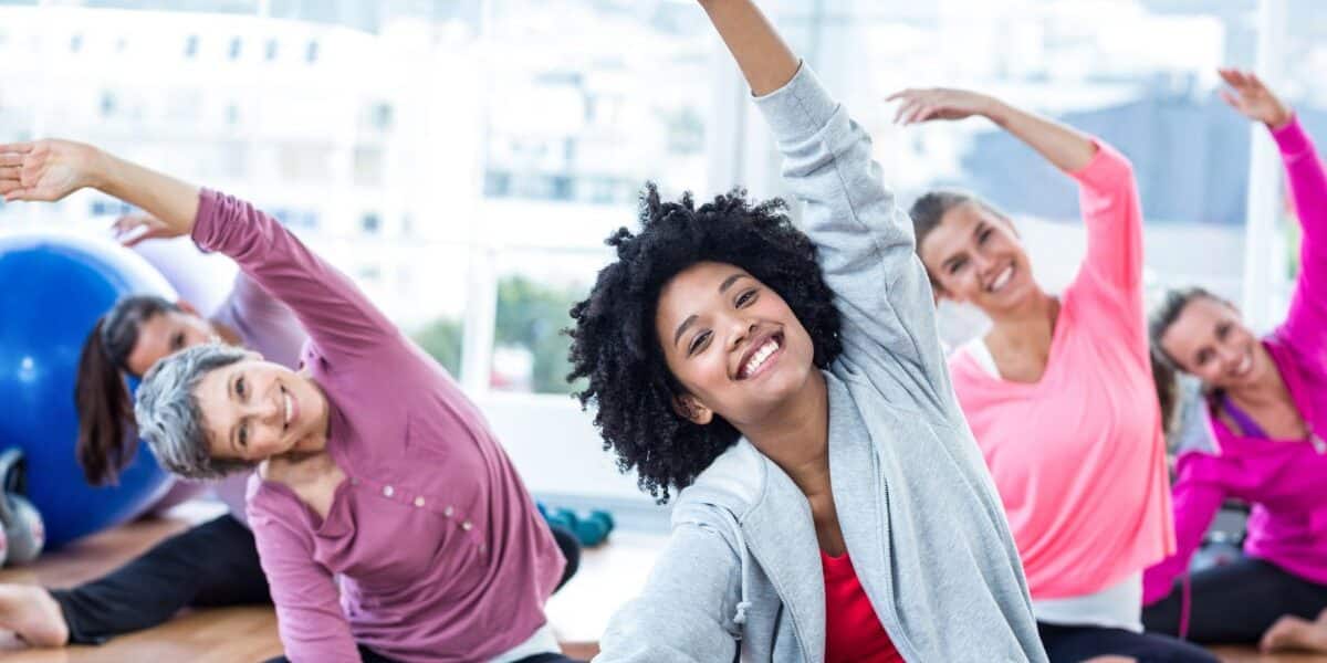 A group of smiling women of various ages sit on yoga mats in a bright studio, stretching their arms overhead to one side. They wear comfortable workout clothes and appear relaxed and happy, engaging in a group fitness or yoga class.