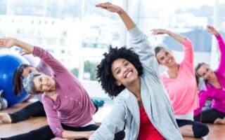 A group of smiling women of various ages sit on yoga mats in a bright studio, stretching their arms overhead to one side. They wear comfortable workout clothes and appear relaxed and happy, engaging in a group fitness or yoga class.