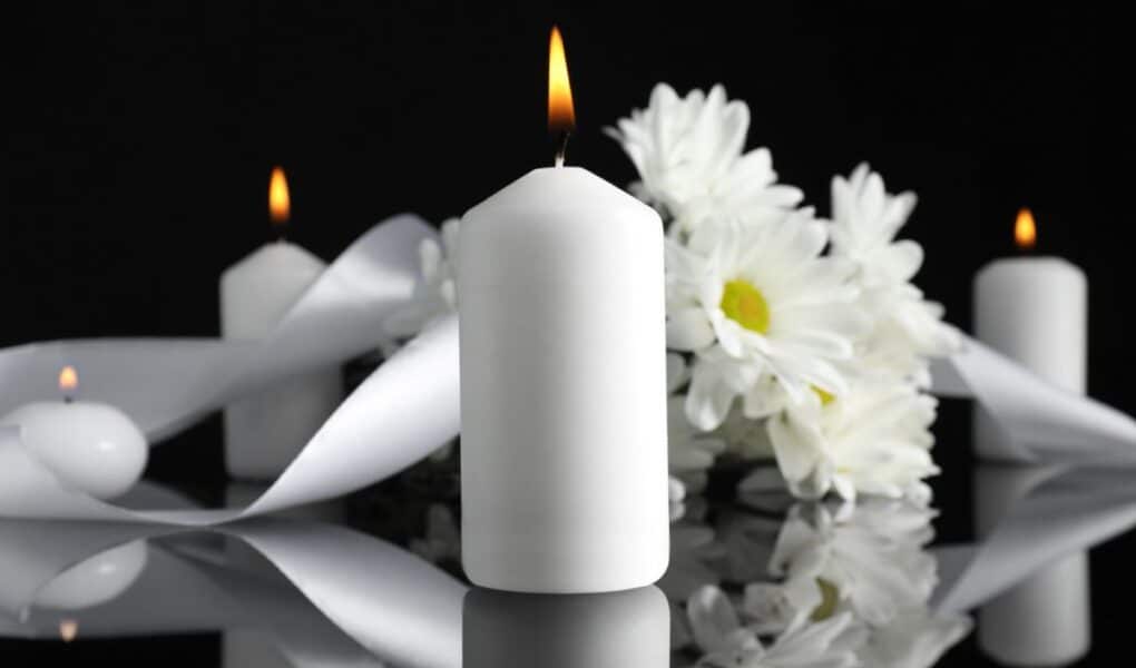 A lit white pillar candle stands on a reflective surface, surrounded by white daisies and a white ribbon. More unlit candles and flowers are softly blurred in the background, all set against a black backdrop. The scene evokes calm and remembrance.