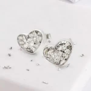 A pair of heart-shaped silver stud earrings with a textured, sparkling surface, resting on a white surface. Small scattered silver beads or crystals are placed around the earrings, enhancing their delicate and elegant appearance.