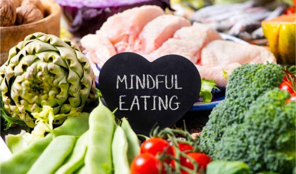 A heart-shaped chalkboard with "MINDFUL EATING" written on it sits among assorted fresh foods, including green beans, cherry tomatoes, broccoli, artichoke, raw chicken, cabbage, and other colourful vegetables.