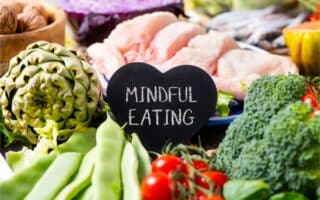 A heart-shaped chalkboard with "MINDFUL EATING" written on it sits among assorted fresh foods, including green beans, cherry tomatoes, broccoli, artichoke, raw chicken, cabbage, and other colourful vegetables.