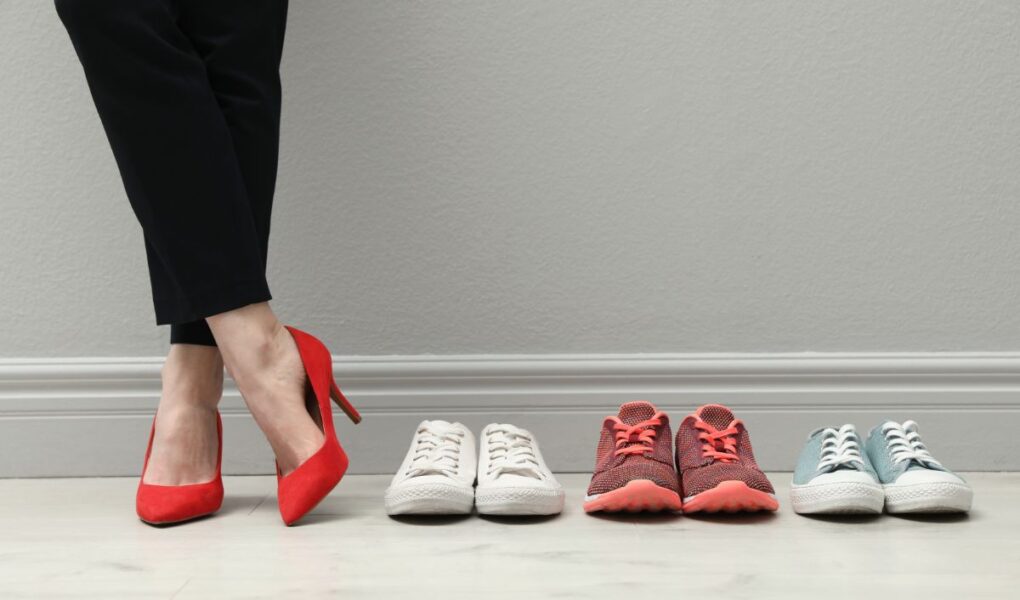 A person wearing black trousers and red high heels stands next to three pairs of trainers—white, red with red laces, and light blue with white laces—all lined up neatly on a light wooden floor against a light grey wall.
