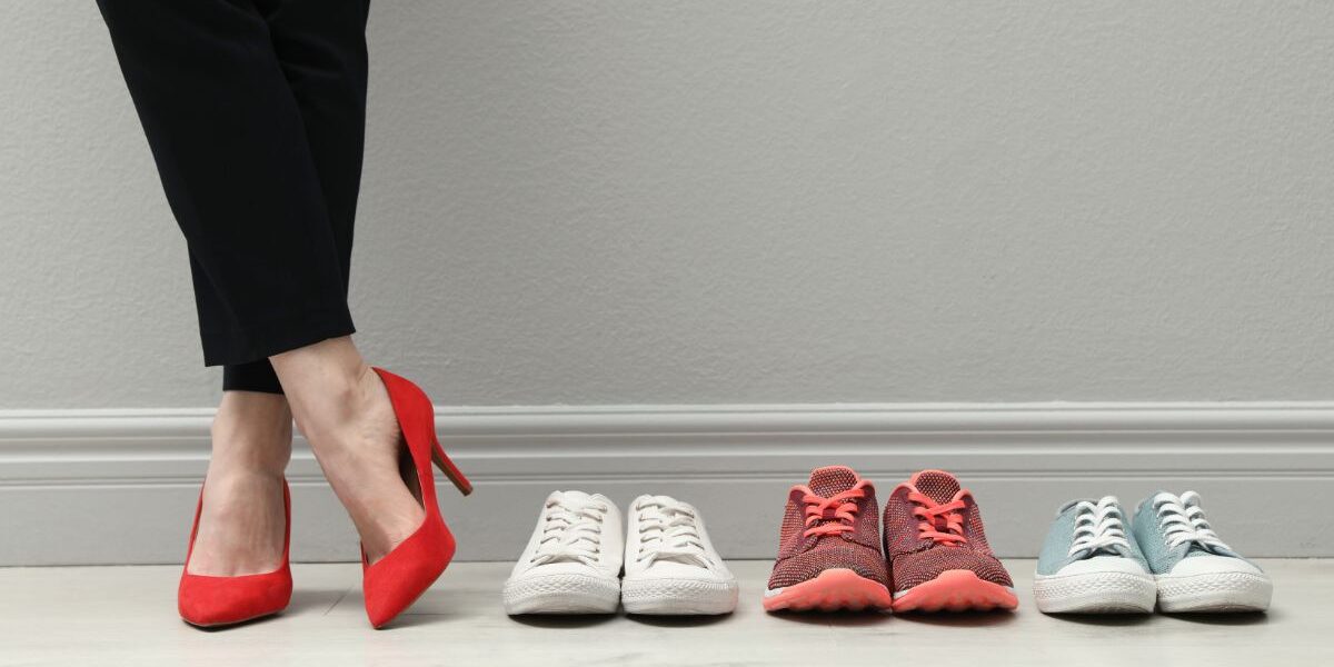 A person wearing black trousers and red high heels stands next to three pairs of trainers—white, red with red laces, and light blue with white laces—all lined up neatly on a light wooden floor against a light grey wall.
