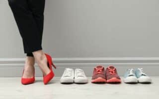 A person wearing black trousers and red high heels stands next to three pairs of trainers—white, red with red laces, and light blue with white laces—all lined up neatly on a light wooden floor against a light grey wall.