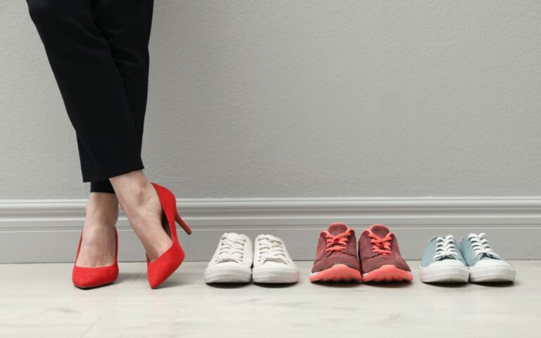 A person wearing black trousers and red high heels stands next to three pairs of trainers—white, red with red laces, and light blue with white laces—all lined up neatly on a light wooden floor against a light grey wall.