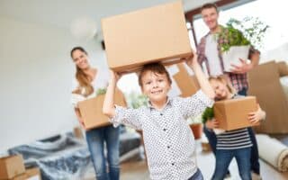 A smiling boy holds a cardboard box on his head while a smiling woman, man, and another child carry boxes and a potted plant in a bright, unpacked living room filled with more boxes and covered furniture, suggesting a family moving into a new home.