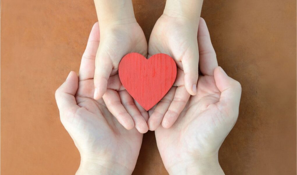 Two adult hands gently cup two smaller child hands, which hold a red wooden heart. The image is taken from above on a brown surface, symbolising care, love, and protection across generations.