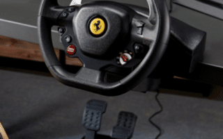 A close-up of a black Ferrari-branded racing game steering wheel mounted to a desk, with paddle shifters and control buttons. Below the desk are two foot pedals, connected to the wheel by a cable, on a grey floor.