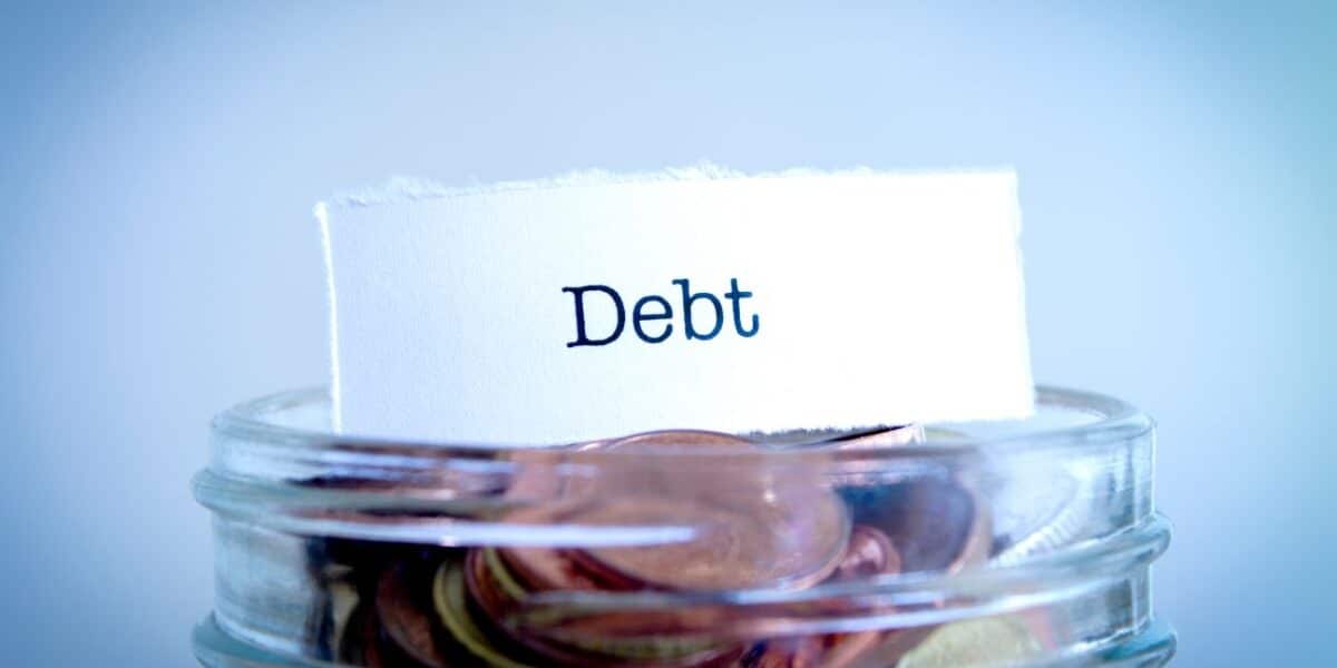A glass jar filled with coins has a white piece of paper sticking out. The paper has the word “Debt” printed in bold, black letters, symbolising financial debt or saving money to pay off debts. The background is softly blurred in blue tones.