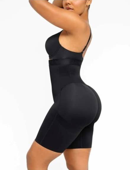 A woman poses in a black high-waisted shaping bodysuit with shorts. The garment fits snugly, smoothing her torso, waist, and thighs. She stands sideways, one arm raised to her head, against a plain white background. Her face is partially visible.