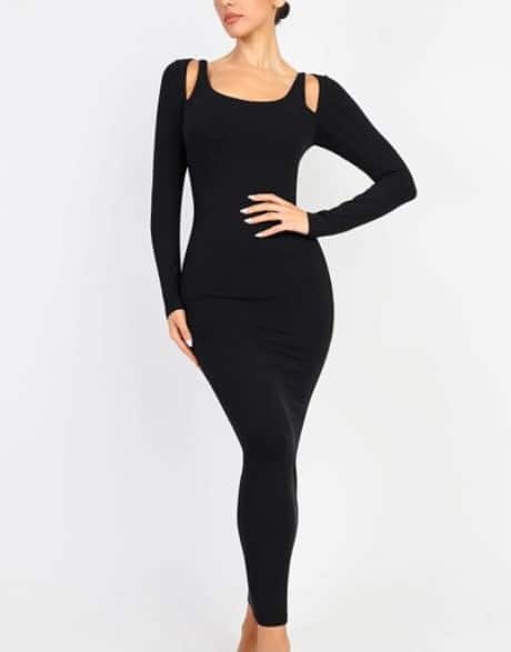 A woman poses in a fitted, long-sleeve, ankle-length black dress with cut-out shoulder details. She stands with one hand on her hip, and her head is slightly turned to the side. The background is plain white.