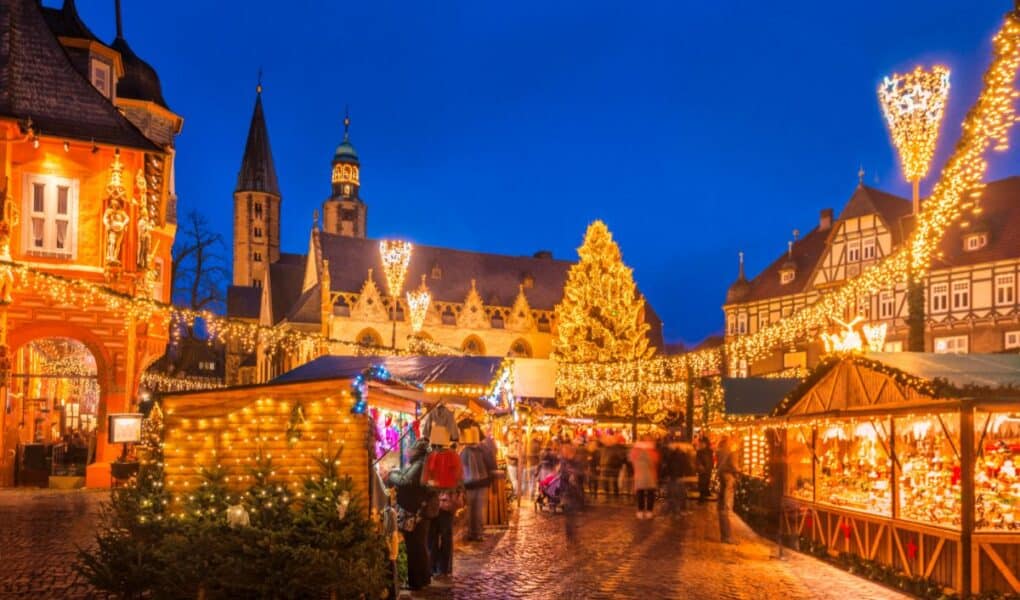 A festive Christmas market at dusk features wooden stalls with lights, decorated trees, and a large illuminated Christmas tree in the centre. Historic buildings surround the square, all outlined in warm fairy lights, beneath a deep blue evening sky.
