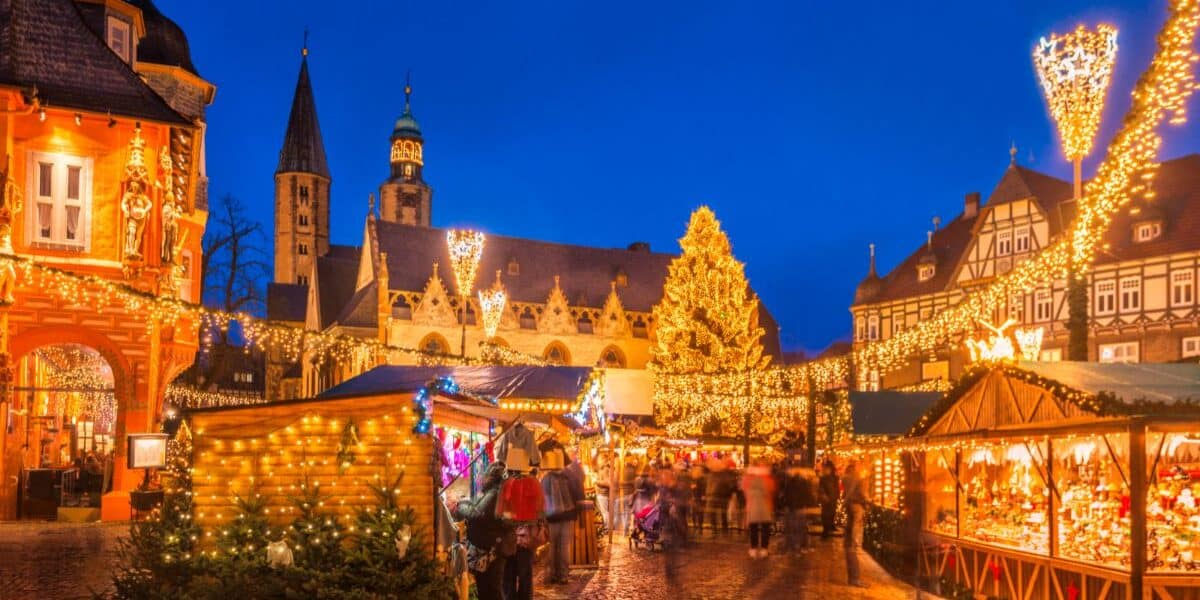 A festive Christmas market at dusk features wooden stalls with lights, decorated trees, and a large illuminated Christmas tree in the centre. Historic buildings surround the square, all outlined in warm fairy lights, beneath a deep blue evening sky.