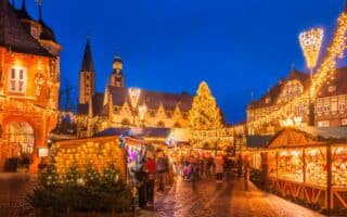 A festive Christmas market at dusk features wooden stalls with lights, decorated trees, and a large illuminated Christmas tree in the centre. Historic buildings surround the square, all outlined in warm fairy lights, beneath a deep blue evening sky.
