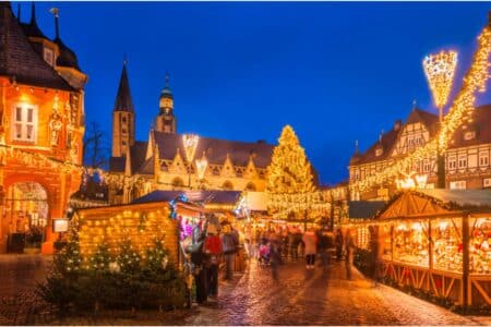 A festive Christmas market at dusk features wooden stalls with lights, decorated trees, and a large illuminated Christmas tree in the centre. Historic buildings surround the square, all outlined in warm fairy lights, beneath a deep blue evening sky.