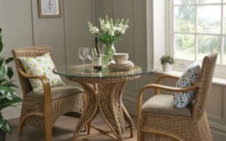 Two wicker chairs with patterned cushions face a round glass-topped table with wooden legs. On the table are white crockery, two wine glasses, a wine bottle, and a vase of white flowers. The scene is by a window with sunlight, wooden floor, and beige panelled walls.
